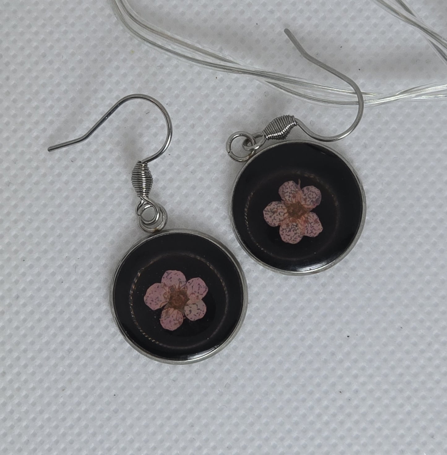 Earrings: single pink Alyssum flowers resting on beds of black acrylic resin, contained in a round silver-coloured stainless steel bezel.