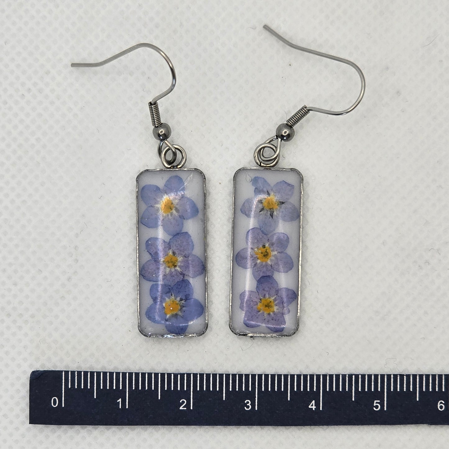 Earrings: Rectangular stainless steel tray-type bezel, filled with white acrylic resin on which three forget-me-not flowers are resting, domed in clear resin,